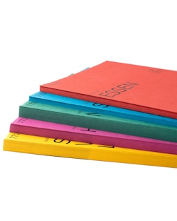 A stack of colorful folders is arranged on a white surface. The folders are in various colors: red, blue, green, pink, and yellow. Each folder has text printed on it.