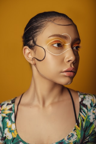 A person with creative makeup featuring yellow and orange eye shadows, highlighted by white lines around the eyes, against a yellow background. The individual wears a top with a floral pattern.