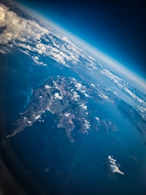 Aerial view capturing a large landmass surrounded by water, with patches of white clouds scattered across the sky. The curvature of the Earth is visible along the horizon, indicating a high altitude perspective.