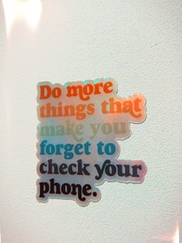 A motivational sticker with the phrase 'Do more things that make you forget to check your phone' is placed on a light-colored, textured background. The text is in various bright colors, including red, blue, green, and black.