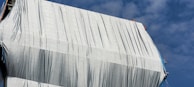 A large structure is covered with a white tarp, with creases and folds visible. In the upper right, two construction workers wearing helmets can be seen at the top edge. The sky is clear with patches of cloud.