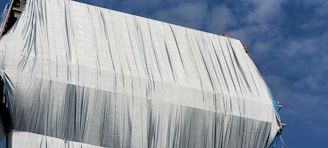 A large structure is covered with a white tarp, with creases and folds visible. In the upper right, two construction workers wearing helmets can be seen at the top edge. The sky is clear with patches of cloud.