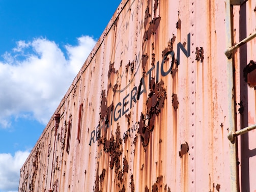 A close-up of a weathered and rusted metal surface, likely of a large container or boxcar. The paint is peeling off, revealing rust underneath. The words 'REFRIGERATION' are partially visible on the side. The sky is bright blue with some fluffy white clouds.