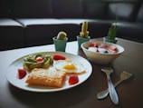 A breakfast setup on a table featuring a plate with a fried egg, slices of avocado, a pastry, and strawberries. Beside it, a bowl is filled with yogurt topped with strawberries and blueberries. Small potted cacti are arranged in the background, and utensils are placed on the table.