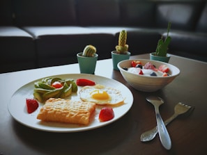 A breakfast setup on a table featuring a plate with a fried egg, slices of avocado, a pastry, and strawberries. Beside it, a bowl is filled with yogurt topped with strawberries and blueberries. Small potted cacti are arranged in the background, and utensils are placed on the table.