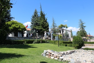 A well-maintained garden area with lush green grass and a variety of trees including pine trees. A stone path or small wall is visible in the foreground. There is a two-story building with a white facade and red-tiled roof in the background, partially obscured by trees and bushes. A flag with red and white colors is prominently displayed near the building. The sky is bright blue with a few scattered clouds, creating a clear and pleasant outdoor atmosphere.