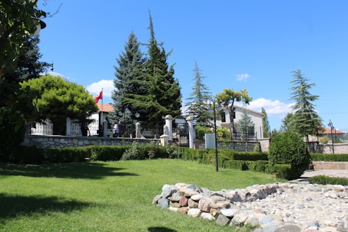 A well-maintained garden area with lush green grass and a variety of trees including pine trees. A stone path or small wall is visible in the foreground. There is a two-story building with a white facade and red-tiled roof in the background, partially obscured by trees and bushes. A flag with red and white colors is prominently displayed near the building. The sky is bright blue with a few scattered clouds, creating a clear and pleasant outdoor atmosphere.