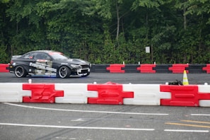 A black car with various sponsor logos is racing on a track surrounded by green trees. The track is lined with red and white barricades. The focus is on the car moving at speed, possibly during a drifting event, with a cone and some barriers visible in the foreground.