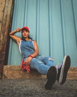 A person is wearing a red cap, blue denim overalls, a sleeveless white shirt, and black sneakers. They are leaning against a corrugated metal wall with one leg extended forward and the other leg bent. The scene features some rusted metal and a rough ground surface.