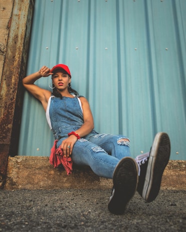 A person is wearing a red cap, blue denim overalls, a sleeveless white shirt, and black sneakers. They are leaning against a corrugated metal wall with one leg extended forward and the other leg bent. The scene features some rusted metal and a rough ground surface.