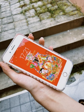 A hand holds a smartphone displaying a colorful and vivid illustration with traditional Chinese elements against a red background. The phone's screen shows an image of festive characters and symbols, possibly related to Chinese culture or celebrations. The phone's time reads 17:21.