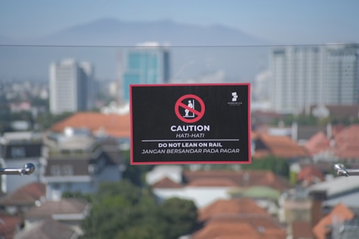 A signboard with a warning message is placed against the backdrop of a cityscape. The sign features a no leaning graphic and the text in both English and Indonesian cautioning people not to lean on the rail.