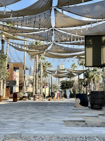 A shaded outdoor area featuring fabric canopies suspended overhead, adorned with string lights. There are palm trees and a couple of barrels positioned randomly. The walkway is paved with stone tiles, and the background shows modern buildings and skyscrapers. A few people are seen in the distance, engaging in casual activities.