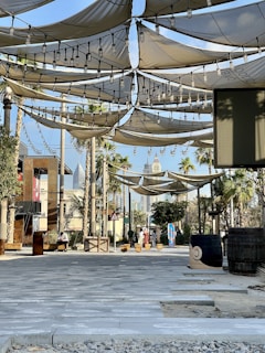 A shaded outdoor area featuring fabric canopies suspended overhead, adorned with string lights. There are palm trees and a couple of barrels positioned randomly. The walkway is paved with stone tiles, and the background shows modern buildings and skyscrapers. A few people are seen in the distance, engaging in casual activities.