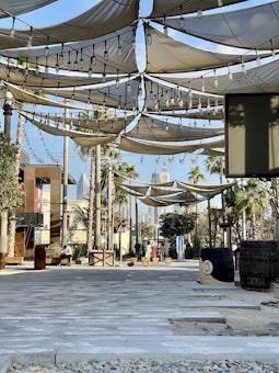 A shaded outdoor area featuring fabric canopies suspended overhead, adorned with string lights. There are palm trees and a couple of barrels positioned randomly. The walkway is paved with stone tiles, and the background shows modern buildings and skyscrapers. A few people are seen in the distance, engaging in casual activities.