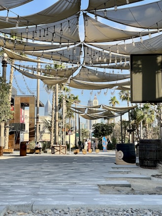 A shaded outdoor area featuring fabric canopies suspended overhead, adorned with string lights. There are palm trees and a couple of barrels positioned randomly. The walkway is paved with stone tiles, and the background shows modern buildings and skyscrapers. A few people are seen in the distance, engaging in casual activities.