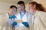 Three scientists in lab coats are engaged in examining a tablet device. Two of them are wearing safety glasses and all are wearing blue gloves. They appear to be focused and in discussion, possibly analyzing data or conducting research.