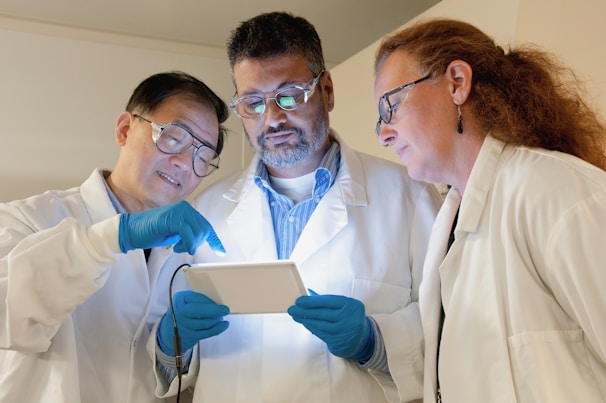 Three scientists in lab coats are engaged in examining a tablet device. Two of them are wearing safety glasses and all are wearing blue gloves. They appear to be focused and in discussion, possibly analyzing data or conducting research.