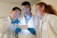 Three scientists in lab coats are engaged in examining a tablet device. Two of them are wearing safety glasses and all are wearing blue gloves. They appear to be focused and in discussion, possibly analyzing data or conducting research.