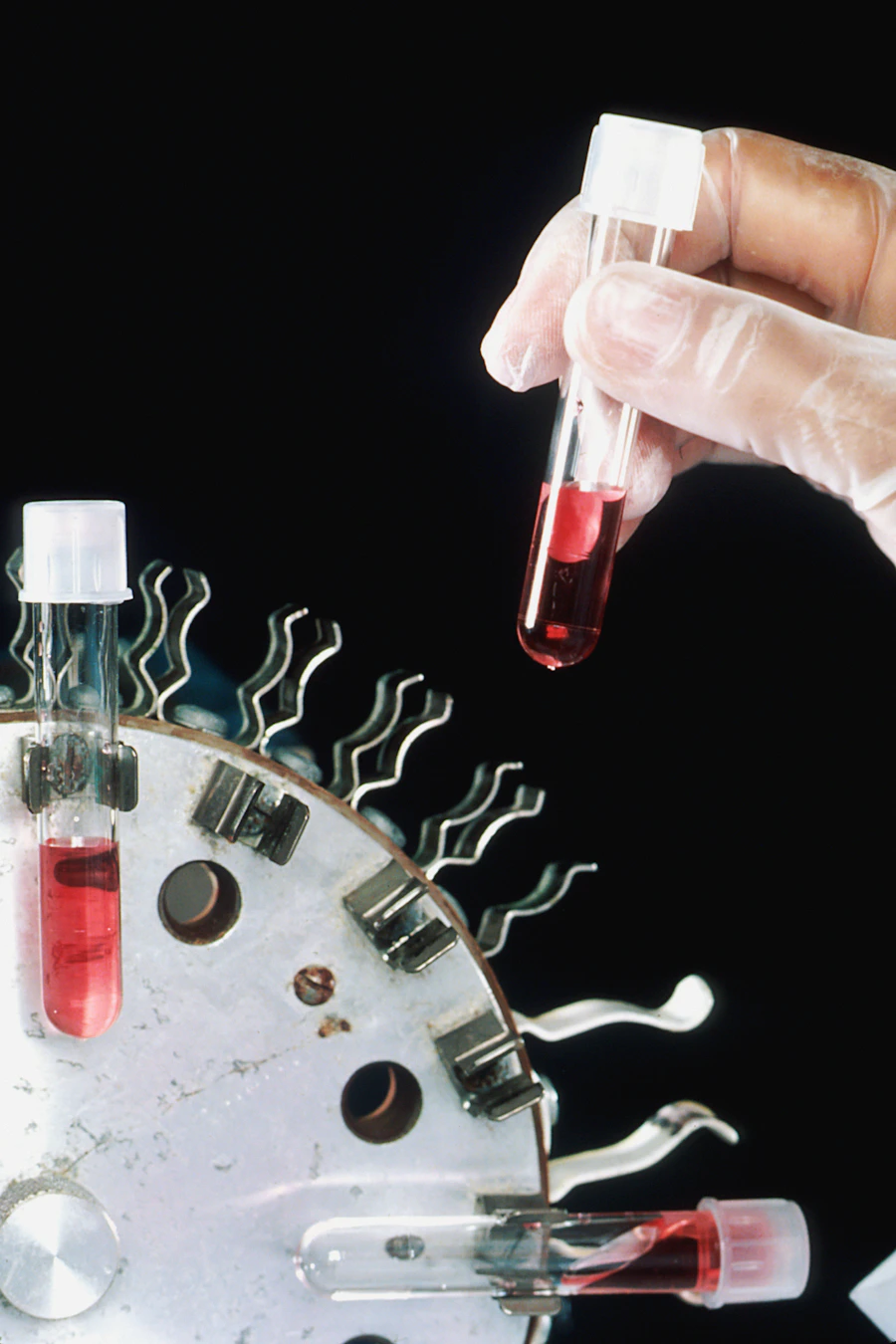 A gloved hand holding a laboratory tube beside laboratory equipment