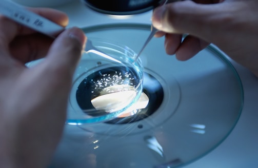 Hands are carefully manipulating a small dish under a microscope. The scene appears to be in a scientific or laboratory setting, possibly involving small particles or samples.