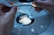 Hands are carefully manipulating a small dish under a microscope. The scene appears to be in a scientific or laboratory setting, possibly involving small particles or samples.