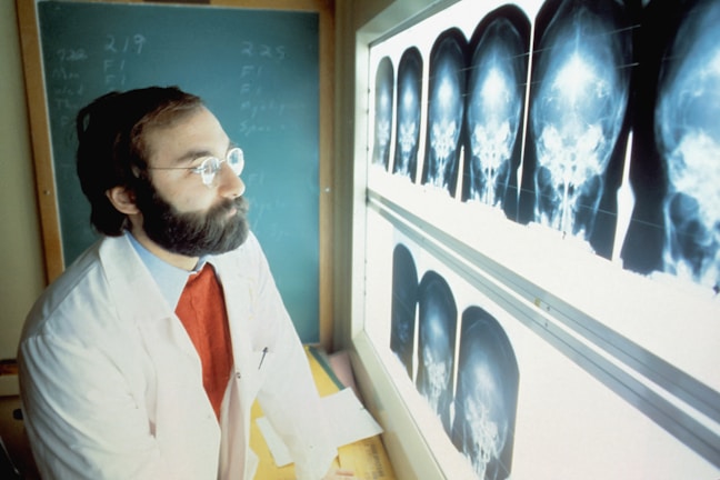 A man with a beard and glasses, wearing a white coat and red sweater, is attentively examining a series of cranial X-ray images displayed on a lightbox. A chalkboard with writing is visible in the background.