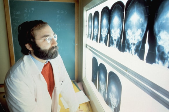 A man with a beard and glasses, wearing a white coat and red sweater, is attentively examining a series of cranial X-ray images displayed on a lightbox. A chalkboard with writing is visible in the background.