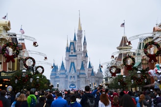 A large, majestic castle with blue and gold spires takes the center of the image, surrounded by festive decorations and a large crowd of people. The decorations include wreaths and ornaments, and American flags can be seen atop some buildings. The scene conveys a festive atmosphere.