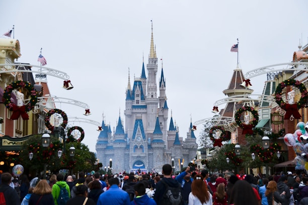A large, majestic castle with blue and gold spires takes the center of the image, surrounded by festive decorations and a large crowd of people. The decorations include wreaths and ornaments, and American flags can be seen atop some buildings. The scene conveys a festive atmosphere.
