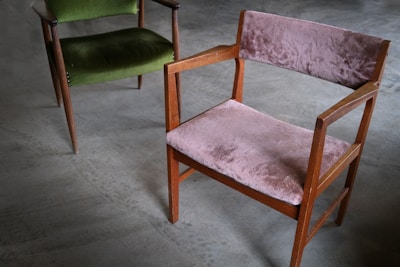 A pair of vintage wooden chairs with plush upholstery are placed on a smooth, grey concrete floor. One chair features a muted pink fabric, while the other has a rich green covering. The chairs have a minimalist, mid-century design with angular armrests and simple frames.
