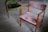 A pair of vintage wooden chairs with plush upholstery are placed on a smooth, grey concrete floor. One chair features a muted pink fabric, while the other has a rich green covering. The chairs have a minimalist, mid-century design with angular armrests and simple frames.