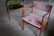 A pair of vintage wooden chairs with plush upholstery are placed on a smooth, grey concrete floor. One chair features a muted pink fabric, while the other has a rich green covering. The chairs have a minimalist, mid-century design with angular armrests and simple frames.