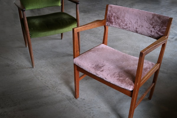 A pair of vintage wooden chairs with plush upholstery are placed on a smooth, grey concrete floor. One chair features a muted pink fabric, while the other has a rich green covering. The chairs have a minimalist, mid-century design with angular armrests and simple frames.