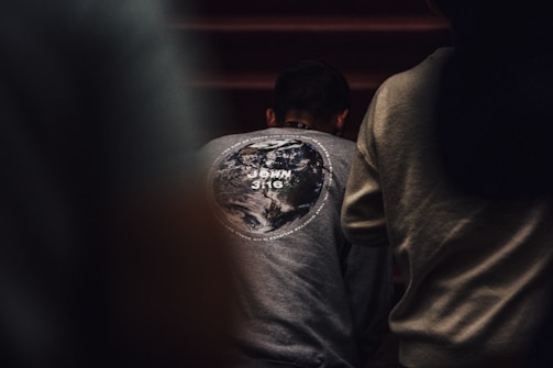 A person is wearing a gray sweatshirt with a design of the Earth on the back, along with the text 'John 3:16.' The surrounding environment is dimly lit, creating a focused atmosphere.