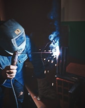 A person wearing a welding mask and gloves is engaged in welding, with bright sparks flying and blue smoke rising. The atmosphere is industrial, with dark surroundings and focused light on the welding point.