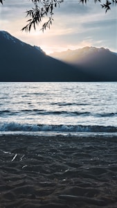A serene lakeside scene at sunset, with gentle waves lapping on the shore. The sky is painted with soft hues of blue and orange as the sun sets behind rugged snow-capped mountains in the background. Silhouetted tree branches frame the top of the image, enhancing the picturesque landscape.