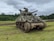 A military tank with a star insignia on the side is positioned on a grassy field. The tank has large tracks and a prominent turret with a long barrel. A person in military attire is standing on top of the tank. The background includes a line of trees under a cloudy sky.