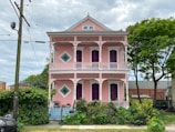 A two-story house painted in pastel pink with ornate white trim and a balcony on each floor. The building features rectangular and diamond-shaped windows, and is surrounded by a lush garden with various shrubs and palm trees. An overcast sky adds a contrast to the vibrant colors of the house.