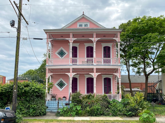 A two-story house painted in pastel pink with ornate white trim and a balcony on each floor. The building features rectangular and diamond-shaped windows, and is surrounded by a lush garden with various shrubs and palm trees. An overcast sky adds a contrast to the vibrant colors of the house.