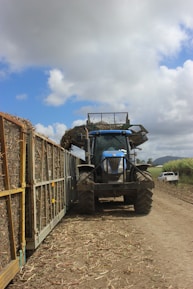 A large blue tractor is positioned on a dirt road next to a series of carts filled with harvested sugarcane. The sky is partly cloudy with patches of blue visible. There are signs of agricultural activity, and a white utility vehicle can be seen in the distance on the road.