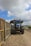 A large blue tractor is positioned on a dirt road next to a series of carts filled with harvested sugarcane. The sky is partly cloudy with patches of blue visible. There are signs of agricultural activity, and a white utility vehicle can be seen in the distance on the road.