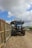 A large blue tractor is positioned on a dirt road next to a series of carts filled with harvested sugarcane. The sky is partly cloudy with patches of blue visible. There are signs of agricultural activity, and a white utility vehicle can be seen in the distance on the road.