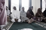 A group of elderly women are seated on a carpeted floor, wearing traditional Muslim attire. Some are in white, while others wear floral and patterned garments. They appear to be in a serene setting, possibly inside a mosque, with windows in the background showing a glimpse of the outside.