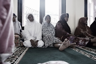 A group of elderly women are seated on a carpeted floor, wearing traditional Muslim attire. Some are in white, while others wear floral and patterned garments. They appear to be in a serene setting, possibly inside a mosque, with windows in the background showing a glimpse of the outside.