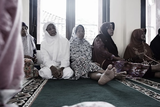 A group of elderly women are seated on a carpeted floor, wearing traditional Muslim attire. Some are in white, while others wear floral and patterned garments. They appear to be in a serene setting, possibly inside a mosque, with windows in the background showing a glimpse of the outside.