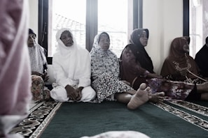 A group of elderly women are seated on a carpeted floor, wearing traditional Muslim attire. Some are in white, while others wear floral and patterned garments. They appear to be in a serene setting, possibly inside a mosque, with windows in the background showing a glimpse of the outside.