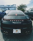 A black Porsche 911 Carrera 4S is parked between two other vehicles in an outdoor setting. The car has a Florida license plate and is positioned in front of a mural depicting ocean scenes with dolphins and stingrays. The background includes palm trees and a partly cloudy sky.