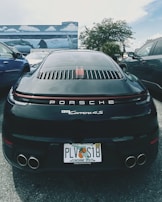 A black Porsche 911 Carrera 4S is parked between two other vehicles in an outdoor setting. The car has a Florida license plate and is positioned in front of a mural depicting ocean scenes with dolphins and stingrays. The background includes palm trees and a partly cloudy sky.
