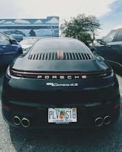 A black Porsche 911 Carrera 4S is parked between two other vehicles in an outdoor setting. The car has a Florida license plate and is positioned in front of a mural depicting ocean scenes with dolphins and stingrays. The background includes palm trees and a partly cloudy sky.